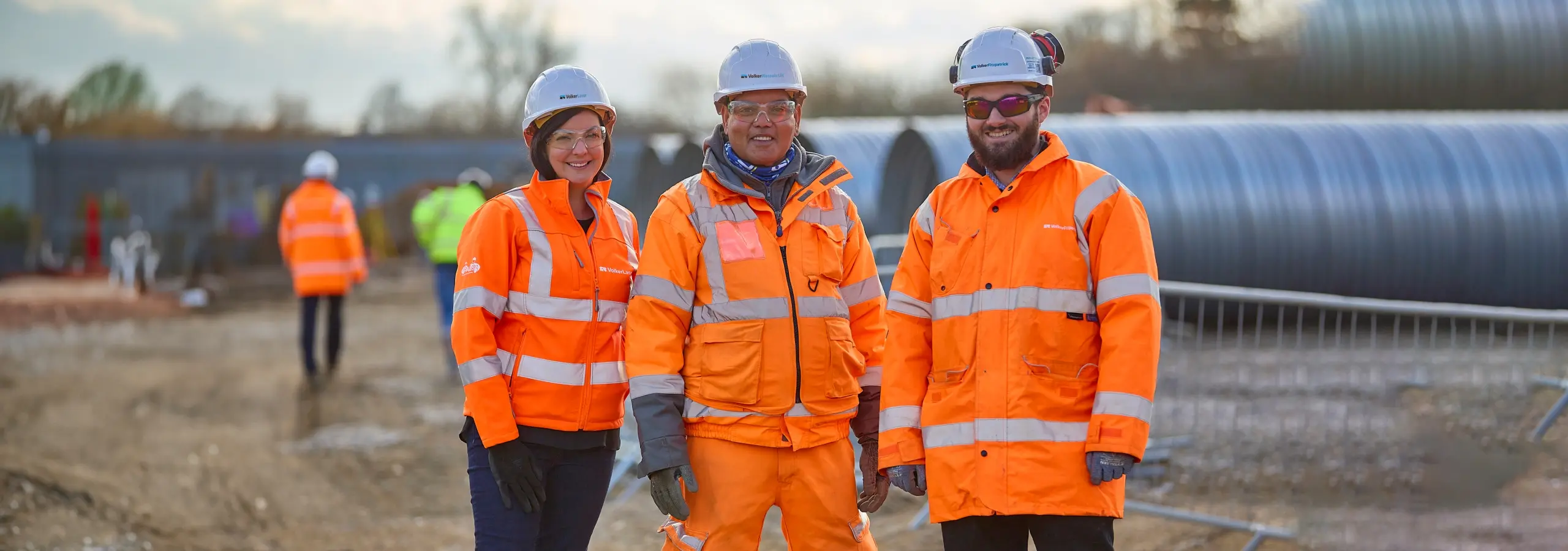 Three VolkerWessels UK construction workers in orange high-visibility PPE and hard hats standing together on an infrastructure site.