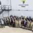 Group of Willmott Dixon staff and military personnel gathered outside stacked modular buildings bearing the Willmott Dixon logo, with two dogs in the foreground.
