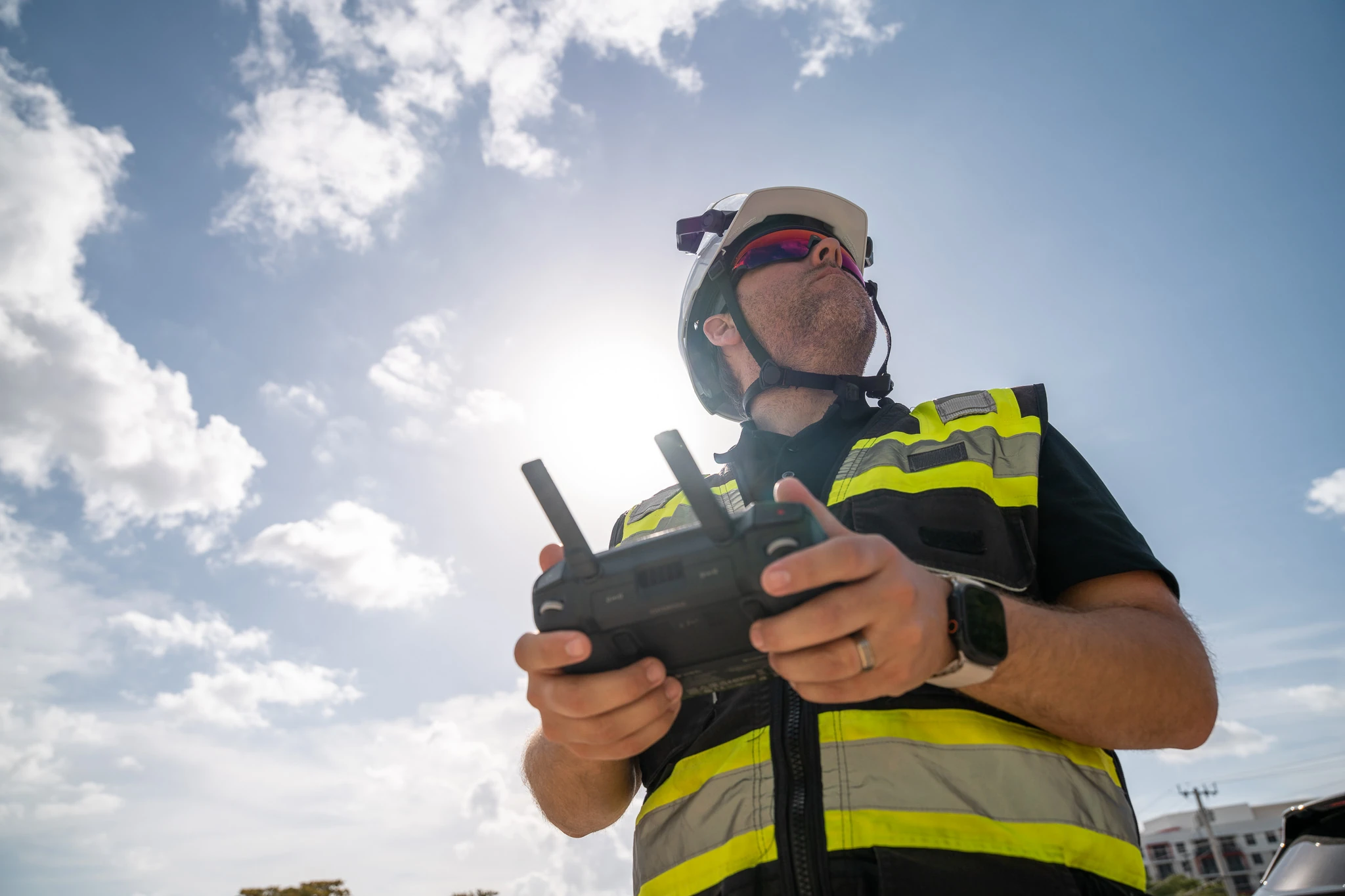 Construction worker wearing a hard hat and high-visibility vest operating a drone controller beneath a bright sky.