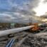 Large underground pipeline being installed using specialist construction equipment on a muddy infrastructure site, with a crane and aircraft visible in the background.