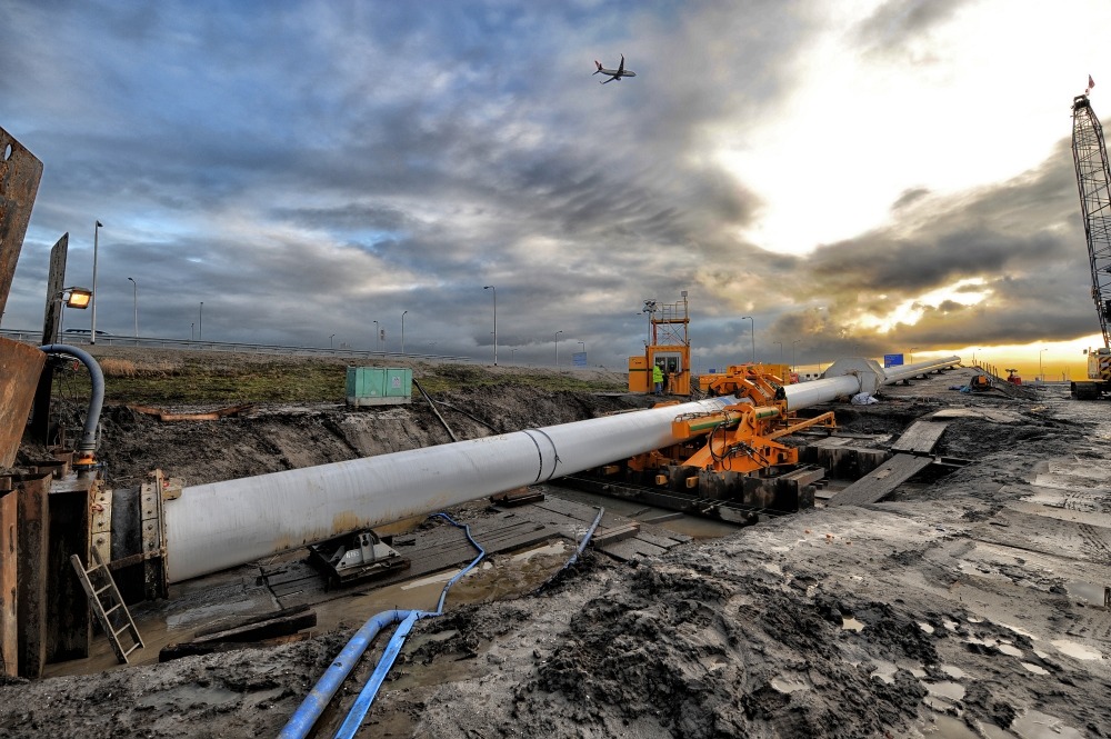 AMAARTENFOTOGRAFIE@UPCMAIL.NL(C)2010 Large underground pipeline being installed using specialist construction equipment on a muddy infrastructure site, with a crane and aircraft visible in the background.